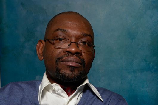 Photograph of John Ogba, Managing Director of GeComm Trade Communication Limited. He wears glasses and is dressed in a white shirt with a sweater over it. Background looks classy. Trade communication at eye level between West Africa, Europe and India.